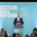 Nigel Farage stood in front of Reform Party UK Sign