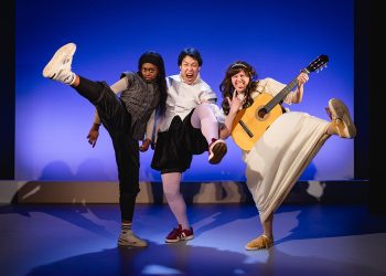 Three performers kick high on stage: a dancer in black, a center dancer leaping, and a guitarist in a white dress wearing yellow shoes against a blue backdrop