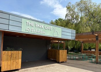 Cafe storefront with a light green sign reading 'THE PARK CAFE' and a closed roller shutter in front. Large wooden planters on wheels flank the entrance.