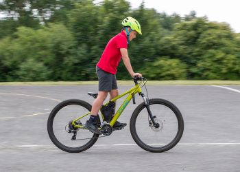 Child wearing a bright yellow helmet rides a lime-green mountain bike on a paved parking lot area with trees in the background, moving to the right.