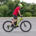 Child wearing a bright yellow helmet rides a lime-green mountain bike on a paved parking lot area with trees in the background, moving to the right.